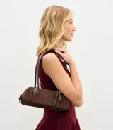 Cartera tipo shoulder bag de tamaño pequeño y formato alargado, confeccionada en material sintético efecto gamuza. Cuenta con asas de hombro estructuradas, cierre superior con cremallera y detalles metálicos decorativos.