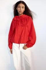 Chaqueta corta de nylon color beige, con cuello subido, capucha y cierre frontal con cremallera. Presenta mangas largas con puños elásticos, bolsillos laterales y detalles de cordones ajustables con lazadas en el pecho y el bajo.