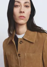 Chaqueta de ante color marrón claro, de diseño corto y recto, con cuello camisero, manga larga, dos bolsillos de parche delanteros y cierre frontal con botones.