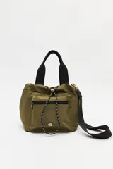Bolso tipo tote/bucket de nylon color verde militar, con asas superiores negras y correa ajustable y desmontable negra. Presenta un bolsillo frontal con cierre y cordón ajustable en la parte superior.