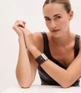 Pulsera ancha de material sintético color marrón oscuro, con diseño de cinta larga y dos tiras metálicas doradas aplicadas en el centro. Cuenta con un cierre de pino ajustable.