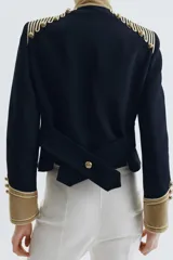 Chaqueta de estilo militar en color negro, con cuello subido y puños en terciopelo color verde oliva. Presenta detalles de alamares dorados en el frente y hombros, bolsillos con solapa y cierre frontal con botones metálicos en relieve.