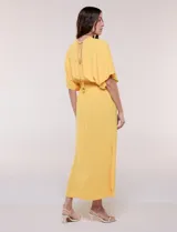 Vestido largo amarillo de corte fluido con escote en V, mangas cortas amplias y apliques de tachas en el escote y puños. Posee cintura elastizada fruncida y falda larga con tajos laterales en el ruedo.