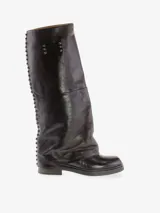 Bota alta de cuero vacuno color marrón oscuro con diseño de polaina integrada. Presenta detalles de tachas metálicas a lo largo de la costura posterior y una placa metálica decorativa en la parte superior de la caña. Posee taco bajo y punta redondeada.