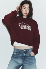 Sudadera de cuello redondo y manga larga, color bordó, con estampado frontal en blanco que dice "Chasing The Sunset's Glow SAN DIEGO 1980 PACIFIC HARBOR".