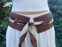 Faja ancha de cuero marrón con costuras decorativas, diseñada para ajustarse en la cintura mediante un pañuelo de seda bicolor en tonos marrón y champagne que se anuda al frente.