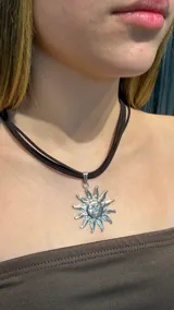 Choker de cordón negro con dije metálico plateado en forma de sol con rostro grabado.