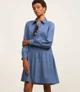 Vestido corto de corte evasé confeccionado en tricolina de algodón, color azul. Presenta cuello camisero, cierre frontal de botones y un panel central con detalle bordado calado. Posee mangas largas con puños abotonados.