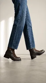 Bota urbana de caña media, confeccionada en una combinación de cuero gastado y gamuza. Presenta una solapa frontal en gamuza, cierre lateral y una tira decorativa con hebilla metálica en el tobillo. Cuenta con una suela de goma robusta y dentada.