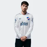 Camiseta de fútbol oficial Umbro del Club Nacional de Football, modelo Home 2026 de manga larga. Diseño en color blanco con cuello tipo polo, detalles en azul y rojo, y estampado de sponsors en el frente y mangas.
