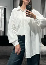 Camisa de corte oversize color marrón, con cuello clásico, cierre frontal de botones y mangas largas con puños ajustables. Presenta un diseño de hombros caídos y un bolsillo frontal en el pecho.