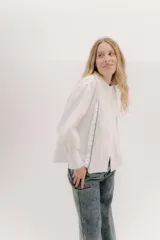 Camisa de algodón blanca con cuello a la base, cierre frontal con botones y mangas largas con puños delicados. Presenta volados verticales y detalles de cintas de terciopelo en contraste a lo largo del frente.