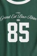 Remera de algodón color verde con cuello y bordes de mangas en color blanco. Presenta un estampado frontal con estilo universitario que incluye el número 85 y la inscripción Grand Est Bas-Rhin.