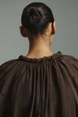 Vestido túnica corto de color marrón con leve brillo, corte oversized, cuello redondo con ribete trenzado y escote en V con tiras finas. Cuenta con mangas globo largas de corte raglán con abertura en los hombros, puños elásticos y cintura fruncida con vuelo en la falda.