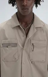 Camisa de manga corta color beige, estilo técnico, confeccionada en microfibra liviana. Cuenta con doble bolsillo frontal, uno con detalle de malla y otro con cierre, y broches en lugar de botones.