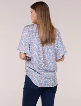 Blusa holgada de poliéster con estampado floral y geométrico en tonos celestes, azules y anaranjados sobre base blanca. Presenta cuello a la base con escote en V y botones frontales, mangas cortas amplias de estilo mariposa y un nudo en el ruedo.