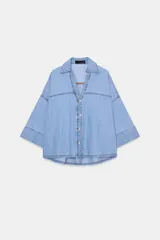 Camisa de denim en tono azul claro, con cuello solapa, escote en pico y cierre frontal con botones. Presenta mangas anchas por debajo del codo y un diseño de bajo asimétrico.