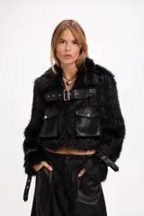 Campera corta de pelo sintético negro con detalles en cuero sintético, incluyendo bolsillos frontales con solapa, cinturón con hebilla metálica en el pecho y correas ajustables en los puños.