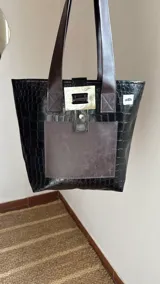 Bolso tote de ecocuero marrón oscuro con textura cocodrilo y un parche frontal de cuero liso marrón claro. Posee asas de hombro de cuero marrón y un broche decorativo blanco en el frente.