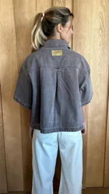 Sobrecamisa de denim soft en tono azul medio, con corte oversize, mangas cortas, cuello clásico y cierre frontal con botones metálicos. Presenta dos bolsillos frontales con solapa y un ruedo redondeado.