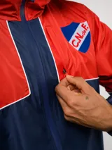 Campera de lluvia Umbro del Club Nacional de Football, con diseño de bloques de color en rojo y azul marino. Presenta capucha, cierre frontal, puños elásticos y detalles de piping blanco. Incluye logo de Umbro y escudo del club en el pecho.