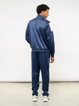 Conjunto deportivo Umbro compuesto por campera de cuello alto con cierre frontal y pantalón de corte recto. La campera presenta bloques de color blanco en los hombros y el logo de la marca bordado en el pecho. Confeccionado en tejido sintético con acabado satinado.