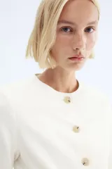 Blazer entallado color blanco roto, con cuello redondo, sin solapas, manga larga y botones frontales de madera. Posee bolsillos de parche delanteros y forro interior.