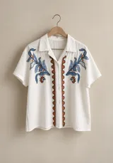 Camisa de manga corta color beige, con cuello camisero, botonadura frontal y bordados decorativos en el frente.