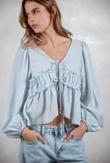 Camisa de tela liviana color celeste, con escote en V y cierre frontal mediante lazos ajustables. Presenta un diseño con volados horizontales en el pecho y mangas largas abullonadas con puños elásticos.