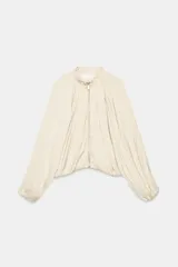 Cazadora satinada de color beige, con cuello alto redondo, mangas largas abullonadas con puños elásticos y cierre frontal de cremallera metálica. Presenta un diseño plisado en todo el cuerpo.