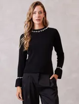 Buzo de tejido soft color negro, con cuello a la base y mangas largas. Presenta apliques de perlas decorativas en el contorno del cuello y en los puños.