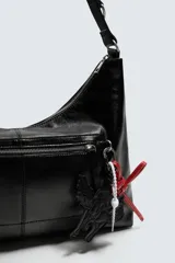 Bolso tipo bandolera de cuero sintético negro con acabado brillante. Presenta un compartimento principal con cierre de cremallera, un bolsillo frontal con cierre y detalle de charms colgantes decorativos. Incluye asa de hombro ajustable.