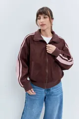 Cazadora de estilo deportivo en color marrón, confeccionada en tejido tipo gamuza. Presenta cierre frontal completo, cuello alto, puños y ruedo acanalados, y franjas laterales en tono rosado. Incluye el logo de la marca bordado en el pecho.