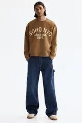 Buzo marrón de algodón con cuello redondo, hombros caídos y diseño estampado en el frente con la inscripción 'SOHO NYC ATHLETICS DEPT.' en color beige. Presenta puños y bajo de canalé elástico.