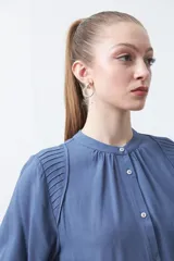 Camisa de color celeste claro con silueta relajada y mangas largas. Presenta un cuello redondo con pequeña abertura frontal y botones, además de un delicado trabajo de tablas en los hombros que aporta textura al diseño.