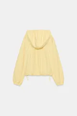 Chaqueta de nylon color amarillo pastel, con cuello subido y capucha. Presenta cierre frontal con cremallera, mangas largas con puños elásticos y detalle de lazadas ajustables en el frente. El bajo cuenta con cordón elástico y stoppers para un ajuste personalizado.