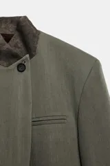 Blazer de corte estructurado en color verde oliva, con cuello subido en tejido a contraste de textura aterciopelada. Presenta cierre frontal con un solo botón, bolsillos con solapa en el delantero y un bolsillo superior tipo ojal.