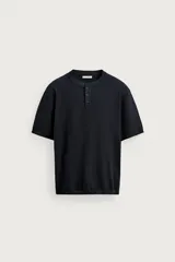Camiseta de punto de algodón color negro, con cuello redondo y cierre frontal parcial de tres botones. Tiene mangas cortas y terminaciones en rib en el cuello, mangas y ruedo.