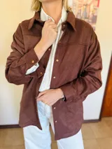 Sobrecamisa de gamuza con cuello clásico, cierre frontal con botones metálicos y mangas largas con puños abotonados. Presenta un diseño estructurado con corte recto y largo a la cadera.