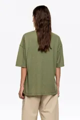 Camiseta de manga corta, corte oversize y escote caja, color verde militar. Presenta un estampado frontal con temática nocturna, incluyendo una luna creciente, árboles y texto en tonos rosados.