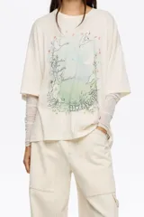Camiseta oversize de algodón y poliéster color crema con cuello redondo y manga corta ancha. Incluye una manga interior ajustada de tul transparente con estampado sutil. Estampado frontal con diseño de paisaje, árboles, aves y texto.