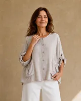 Blusa beige de lino de corte holgado y silueta oversize, con cuello redondo, cierre frontal de botones, bolsillos aplicados grandes y mangas tres cuartos con detalles de perforaciones bordadas en las mangas y el ruedo.