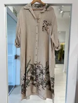 Vestido midi beige de lino, con cierre frontal abotonado, cuello camisero, mangas largas y bolsillos. Presenta un estampado floral en tonos marrones y verdes en la parte inferior, el bolsillo del pecho y el cuello.