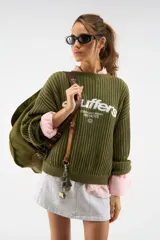Suéter de punto verde oliva de la marca Scuffers, con fit relajado, mangas amplias y texto bordado en blanco en el frente.