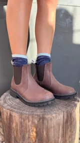 Bota tipo Chelsea de cuero marrón con textura granulada, paneles laterales elásticos en color negro y suela track dentada con costura blanca visible.