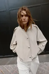 Trench corta de color beige, con cuello solapa, mangas largas con trabillas en los hombros, bolsillos delanteros de vivo y cierre frontal cruzado con botones a presión.