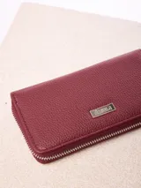 Billetera rectangular de cuero sintético con textura granulada, color bordó. Cuenta con cierre perimetral metálico y placa metálica con el logo de la marca en el frente.