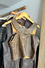Top corto de cuero ovino marrón con efecto veteado, diseño sin mangas con cuello redondo y detalles de pasamanería en la unión de las piezas.