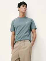 Camiseta blanca de algodón con cuello redondo y mangas cortas. Estampa rectangular con borde negro que contiene la frase "NATURAL ATMOSPHERE" en letras mayúsculas y una imagen de montañas y mar.