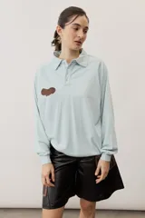 Buzo celeste de manga larga con cuello polo y cierre frontal de botones. Presenta un parche marrón bordado en el pecho.